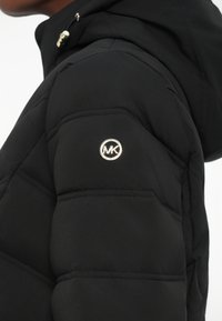 Doudoune noire avec un design matelassé et une capuche. Présente un logo circulaire argenté sur l'épaule gauche. Texture lisse.