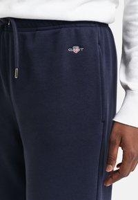 Tumesinised dressipüksid, millel on tikitud GANT logo taskualal, kantud valge pika varrukaga särgiga, näidates inimese kätt küljel.