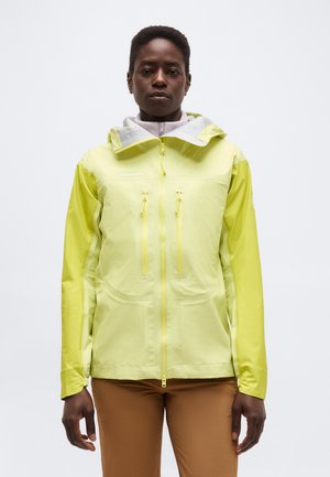 Osoba oblečená v svetložltej kapucňovej bunde so zipsom vpredu a náprsné vrecká, v kombinácii s hnedými nohavicami, stojaca na bielom pozadí.