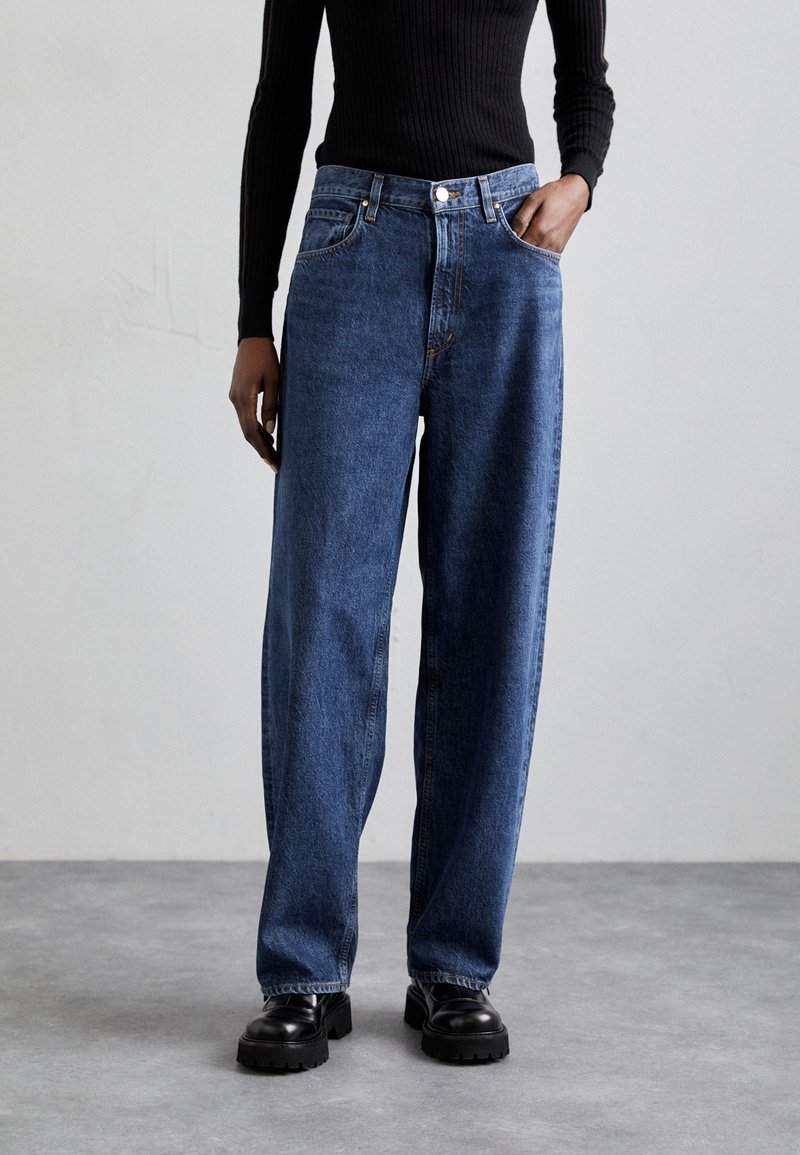 Jean large en denim bleu, avec une taille haute, des poches avant et une fermeture à bouton. Associé à des bottes noires à semelle épaisse.