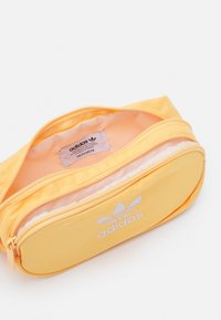 Žlutá taška na zip z odolné tkaniny, se zataveným bílým logem Adidas a hladkým vnitřním podšívkou. Úhlový tvar se šitými detaily.