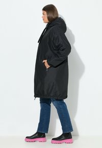 Zwarte waterdichte jas met een capuchon, elastische taille en zijzakken, gecombineerd met blauwe jeans en zwarte laarzen met een roze zool.