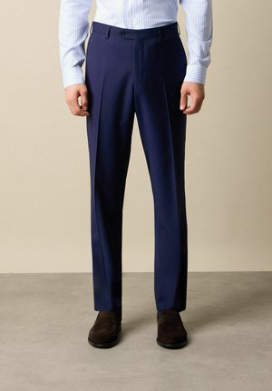 Man met op maat gemaakte marineblauwe nette broek, lichtblauw gestreept overhemd en donkerbruine suède loafers staand op een grijze vloer.