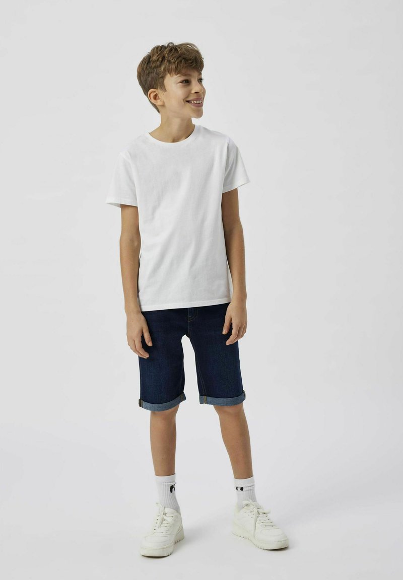 Fehér rövid ujjú póló sötétkék farmer rövidnadrággal, felhajtott szegéllyel, fehér tornacipővel és mintás zoknival viselve.