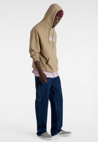Felpa beige con cappuccio e logo bianco, indossata sopra una maglietta rosa chiaro, abbinata a jeans a gamba larga blu scuro e scarpe a scacchi bianco e nero.