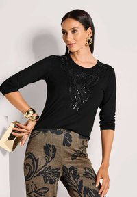 Schwarzes Langarmoberteil mit verziertem Blumenmuster auf der Vorderseite, kombiniert mit dunklen gemusterten Hosen mit großen Blumenmotiven.