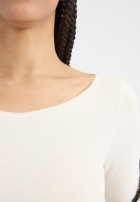 Hvit ribbet topp med bred utringning og glatt tekstur, med en tettsittende design og subtile sømdetaljer langs kantene.