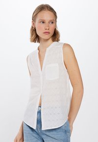 Bílý bezrukávový top s diamantovým vzorem a detaily s výřezem, zapínání na knoflíky vpředu a jednou kapsou na prsou. Vyrobeno z lehkého materiálu.