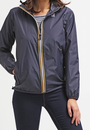 Femme portant une veste coupe-vent bleu marine avec une fermeture éclair rayée et une chemise rayée en dessous, debout avec une main dans la poche.