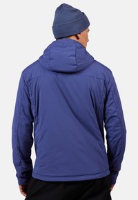 Blaue isolierte Jacke mit Kapuze, die eine glatte Textur aufweist, Nähte entlang der Schultern hat und eine taillierte Passform bietet. Getragen mit einer passenden Mütze.