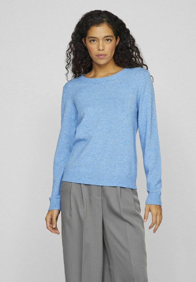 Gaiši zils, adīts džemperis ar apaļu kakla izgriezumu un ribotām piedurknēm. Kombinēts ar pelēkām svītrainām biksēm.