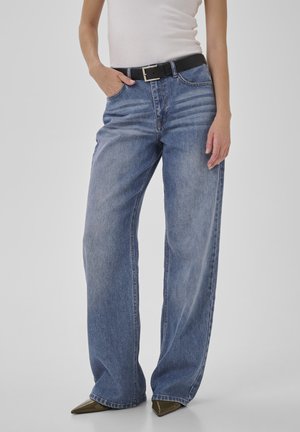 Donna che indossa jeans larghi a vita alta blu, top bianco aderente senza maniche, cintura nera e scarpe verdi oliva a punta.