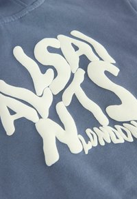 Felpa blu con testo ondulato bianco audace. Tessuto morbido e texturizzato con design a girocollo e dettagli di cucitura visibili.