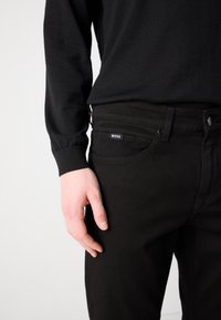 Crne traperice uskog kroja, s branded oznakom na prednjoj džepu i glatkom, teksturiranom završnom obradom. Upotpunjene crnom košuljom dugih rukava.