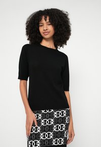 Μαύρο πουλόβερ με κοντά μανίκια και στρογγυλή λαιμόκοψη, φτιαγμένο από μαλακό υλικό. Συνδυασμένο με μπλούζα και φούστα σε ασπρόμαυρο μοτίβο.