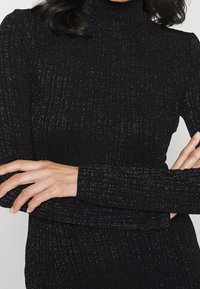 Schwarz gerippter Rollkragenpullover mit metallischen Fäden, langen Ärmeln und taillierter Silhouette. Die Textur ist weich mit einem dezenten Glanz.