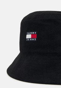 Cappello a secchiello in corduroy nero con un'etichetta ricamata che presenta il testo "TOMMY JEANS" e un design rettangolare rosso, bianco e blu.