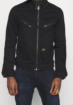 Musta vetoketjulla varustettu takki, jossa on kaksi vinosti aseteltua rintataskua vetoketjuilla ja napilliset hihansuut, puettuna tummien farkkujen päälle.