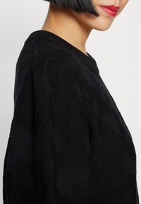Suéter negro de punto con un patrón texturizado, cuello de canalé y un corte holgado, mostrado desde un ángulo lateral, destacando los detalles y la tela.