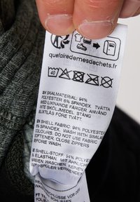 Etichetta con istruzioni per la cura, contenuto del tessuto: 94% poliestere, 6% spandex, in più lingue. Sfondo bianco con testo nero.