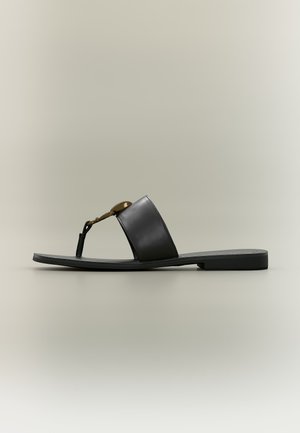 Sandalo nero piatto con fascia larga in pelle, infradito e dettaglio metallico circolare dorato, mostrato di profilo su sfondo neutro.