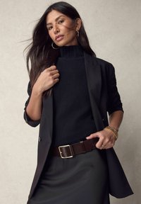 Blazer nero sopra un dolcevita nero, cintrato con una cintura marrone con una fibbia d'oro. Capelli lunghi e bracciali d'oro aggiungono dettagli.