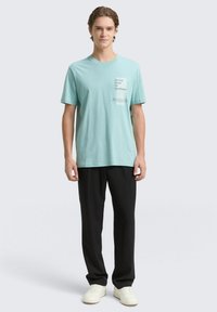 Svetlo modra bombažna majica s kratkimi rokavi in okroglim izrezom, na katero je natisnjen grafični vzorec na prsih. Oblečena z črnimi hlačami in belimi supergami.