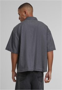 Maglietta grigia a maniche corte in tessuto strutturato, con colletto a punta e spalle scese, vista da dietro, abbinata a jeans scuri.