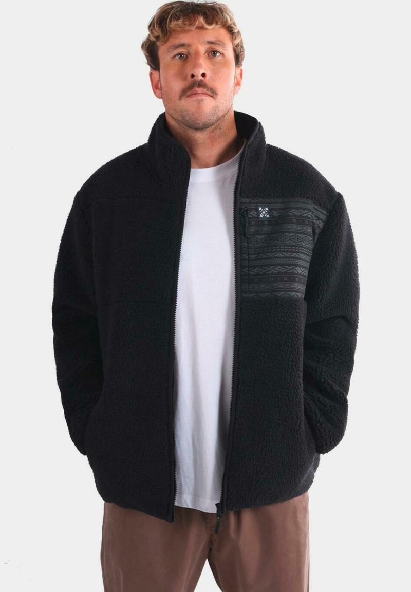 Hombre con cabello rizado castaño claro y bigote, vistiendo una chaqueta de forro polar negra con un bolsillo en el pecho de diseño, camiseta blanca y pantalones marrones, con las manos en los bolsillos.