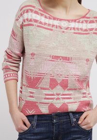Pletený svetr s kulatým výstřihem, který má geometrický vzor v odstínech růžové a béžové. Má dlouhé rukávy a žebrované manžety.