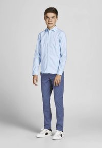 Šviesiai mėlyna marškiniai su sagomis, mėlynos kelnės ir balti sportbačiai juodais akcentais. Paprastas dizainas, prigludusi forma, lygi tekstūra. Stovinčio žmogaus poza.