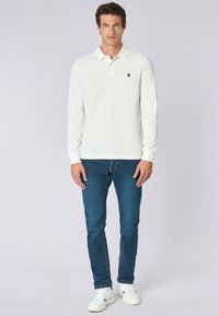 Polo shirt bianco a maniche lunghe con un piccolo logo sul petto sinistro, abbinato a jeans blu e sneakers bianche, posizionato su uno sfondo neutro.