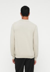 Sweater bege claro, textura tricotada, mangas longas e punhos canelados. Apresenta uma gola clássica e um ajuste ligeiramente solto.