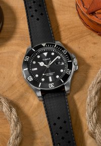 Timex ūdensizturīgs rokas pulkstenis ar melnu ciparnīcu, baltām atzīmēm, datuma displeju un teksturētu melnu gumijas siksniņu uz koka virsmas.