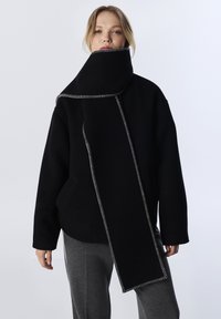 Abrigo negro de gran tamaño con cuello alto y bufanda larga. Presenta costuras blancas en contraste, textura suave y un ajuste relajado.