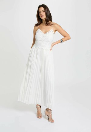 Donna che indossa un vestito bianco in pizzo con gonna a pieghe e sandali argentati con tacco alto, in piedi davanti a uno sfondo semplice.