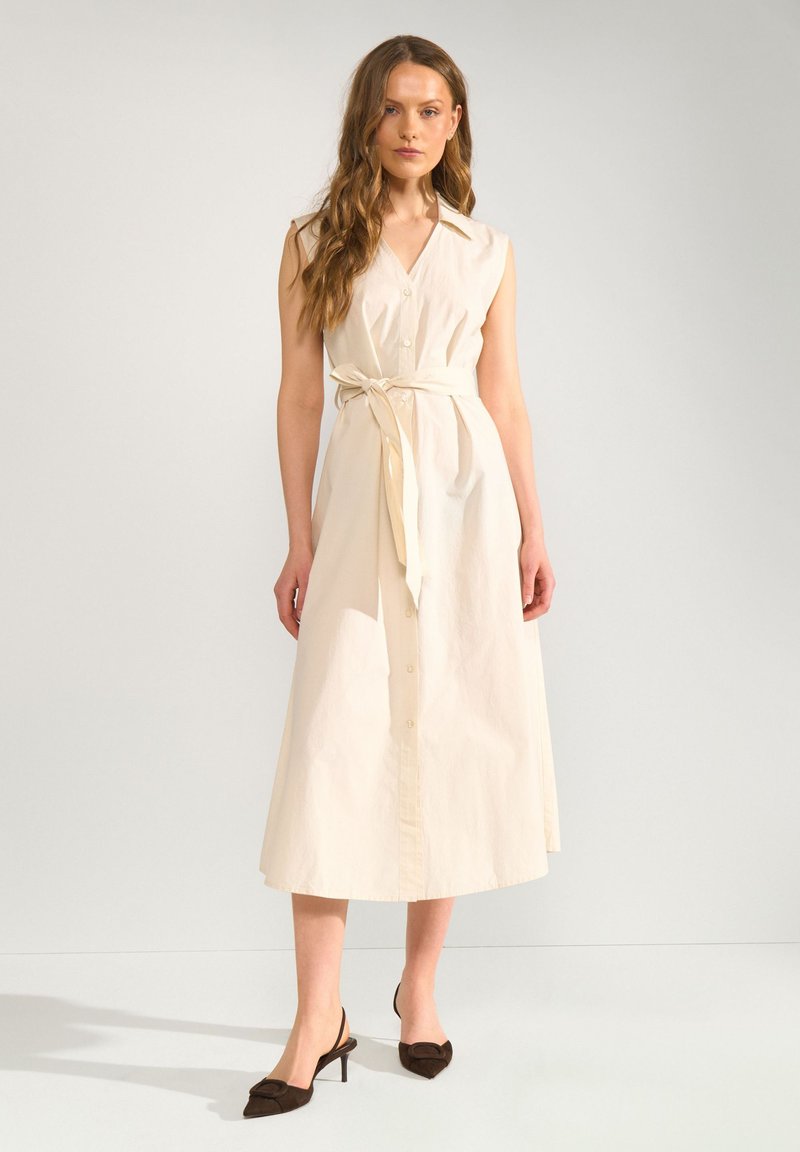 Femme aux longs cheveux ondulés portant une robe midi beige sans manches avec une ceinture nouée à la taille et des talons noirs pointus, debout sur un sol blanc.