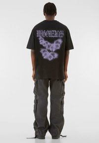 Tricou din bumbac negru, cu un grafic mare de fluture fantomă și text pe spate. Asortat cu pantaloni cargo lărgi de culoare gri închis.
