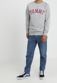 Sudadera gris con el texto "TOMMY" en arco rojo, combinada con jeans azules y zapatillas negras con cordones blancos y detalles azules.
