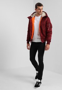Jaqueta bomber bordeaux com forro laranja e gola de pelúcia sintética; bolsos com fecho zip; usada sobre um sweatshirt branco e jeans pretos. Ténis pretos.