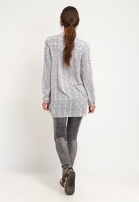 Chemise à manches longues avec un motif géométrique gris et blanc, tissu léger, coupe décontractée, avec des fentes latérales et un ourlet arrondi.