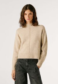 Beige, kort stickad tröja med hög hals, ribbad nederkant och vertikal sömdekoration. Matchad med mörka, löst sittande jeans.
