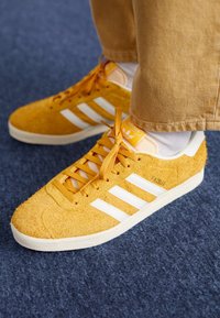 Žluté semišové tenisky s bílým designem tří pruhů a sladěnými tkaničkami. Mají zesílenou špičku a texturovanou podrážku.