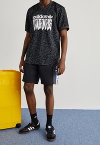 Camiseta gráfica negra de adidas con un patrón abstracto, combinada con pantalones cortos negros con un acento azul y blanco, y zapatillas negras con rayas blancas.