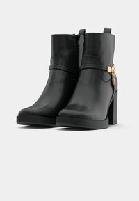 Crne gležnjače s sjajnom površinom, blok peta, bočni patentni zatvarač i dekorativna traka s zlatnim detaljima. Teksturirana potplata za bolje prianjanje.