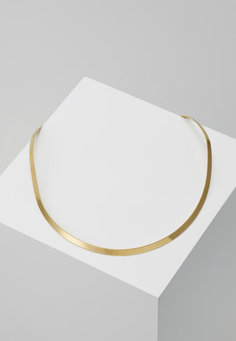 Goldfarbene Halskette mit einem schlanken, flachen Design und einer strukturierten Oberfläche, die auf einem weißen Podest vor einem grauen Hintergrund ruht.