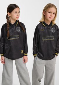 Svart långärmad polo med en texturerad mönster, gula accenter och logotyp, tillsammans med grå vida byxor.