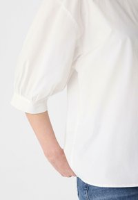 Biała bawełniana koszula z bufiastymi rękawami i mankietami. Gładka tekstura, boczne rozpórki i luźny krój. Stylizowana z niebieskimi dżinsami.