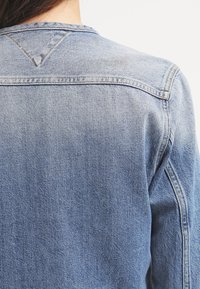 Ljusblå jeansjacka med en klassisk design, synliga sömmdetaljer och en triangulär ok på baksidan. Mjuk textur.