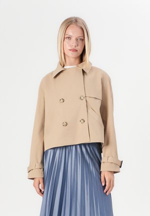 Veste beige courte à double boutonnage avec un col, deux poches avant et des accents de boutons, associée à une jupe plissée bleue.